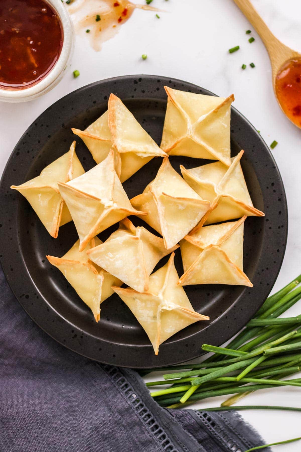 black plate with a pile of cream cheese wontons, bowl of chili dipping sauce, chives