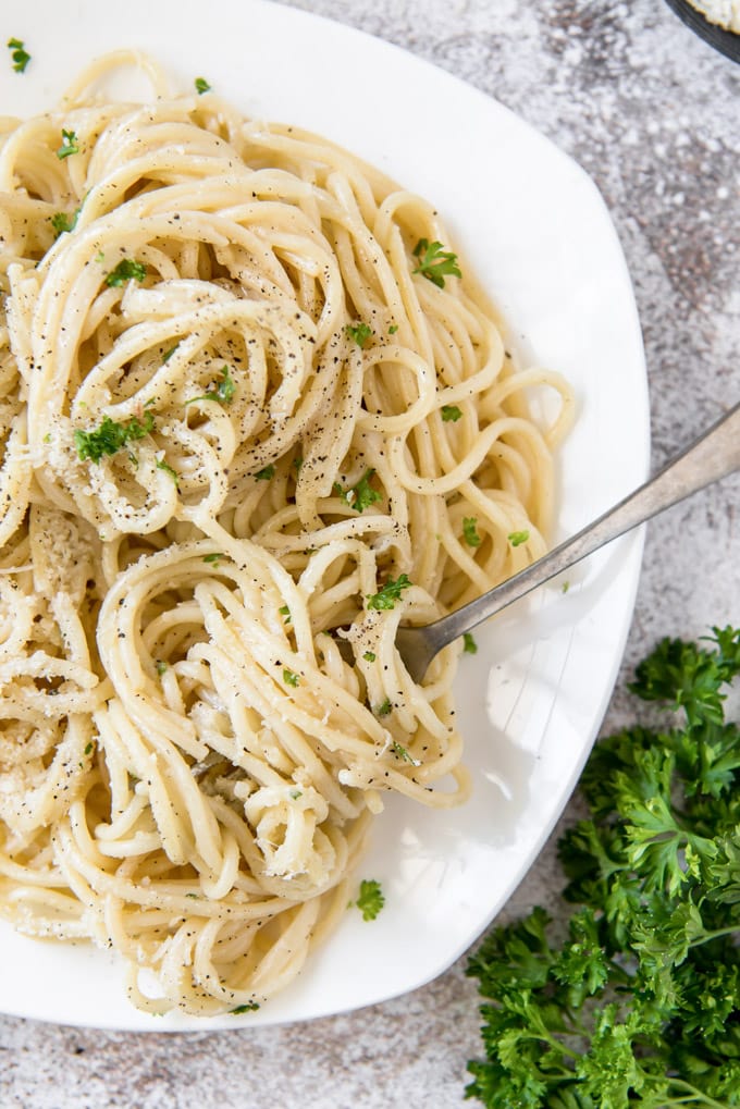 a white plate with spaghetti pasta with cheese, black pepper and parsley. A fork twirls the pasta off to the side.