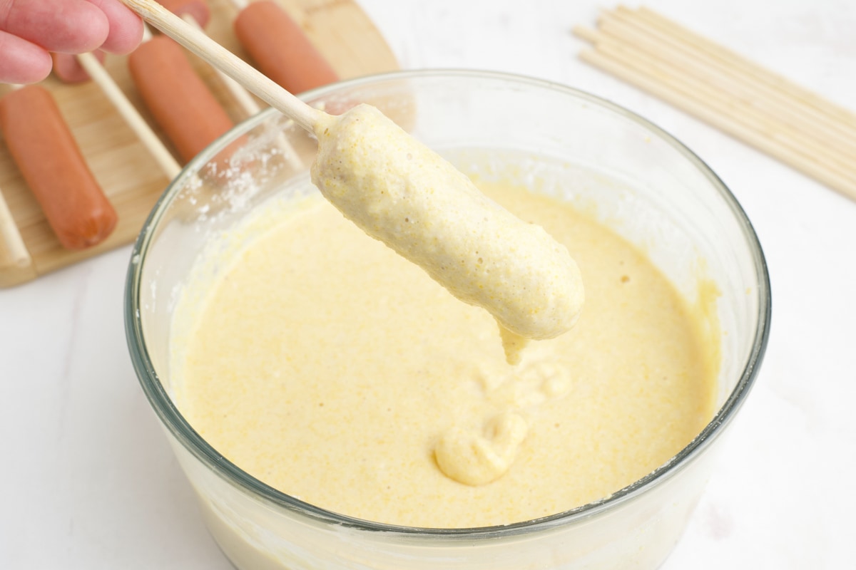 Corn dog dipped in batter from a clear glass bowl.