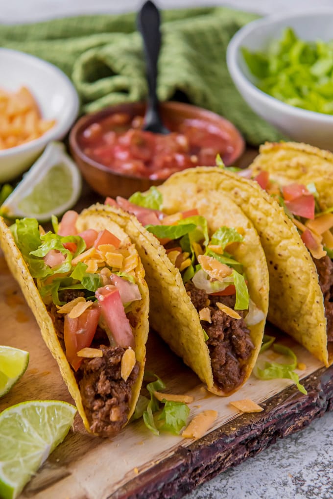 Ground Beef Tacos setup on a platter surrounded by bowls of salsa, cheese, guacamole and lime wedges.