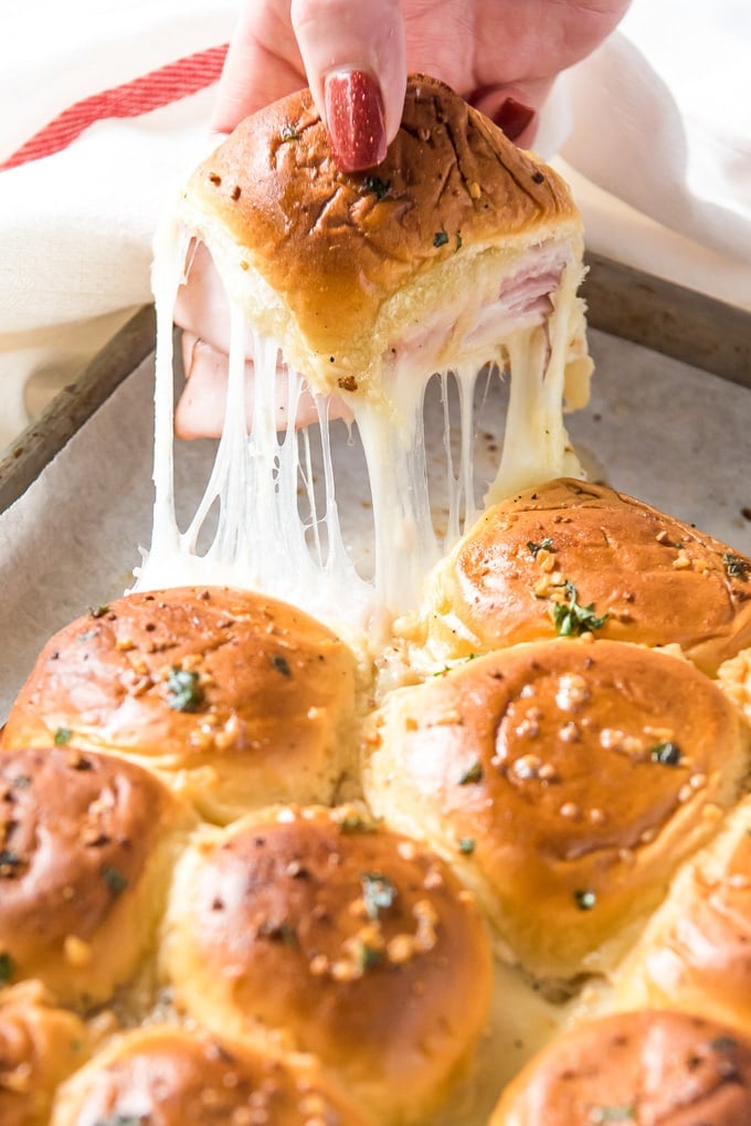 a hand reaching in and pulling out a ham and cheese slider with strings of melted cheese