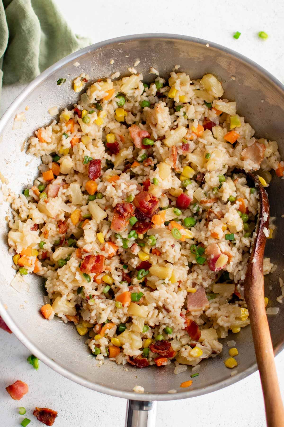 Fried rice with bacon and pineapple and green onions in a silver wok with a wooden spoon.