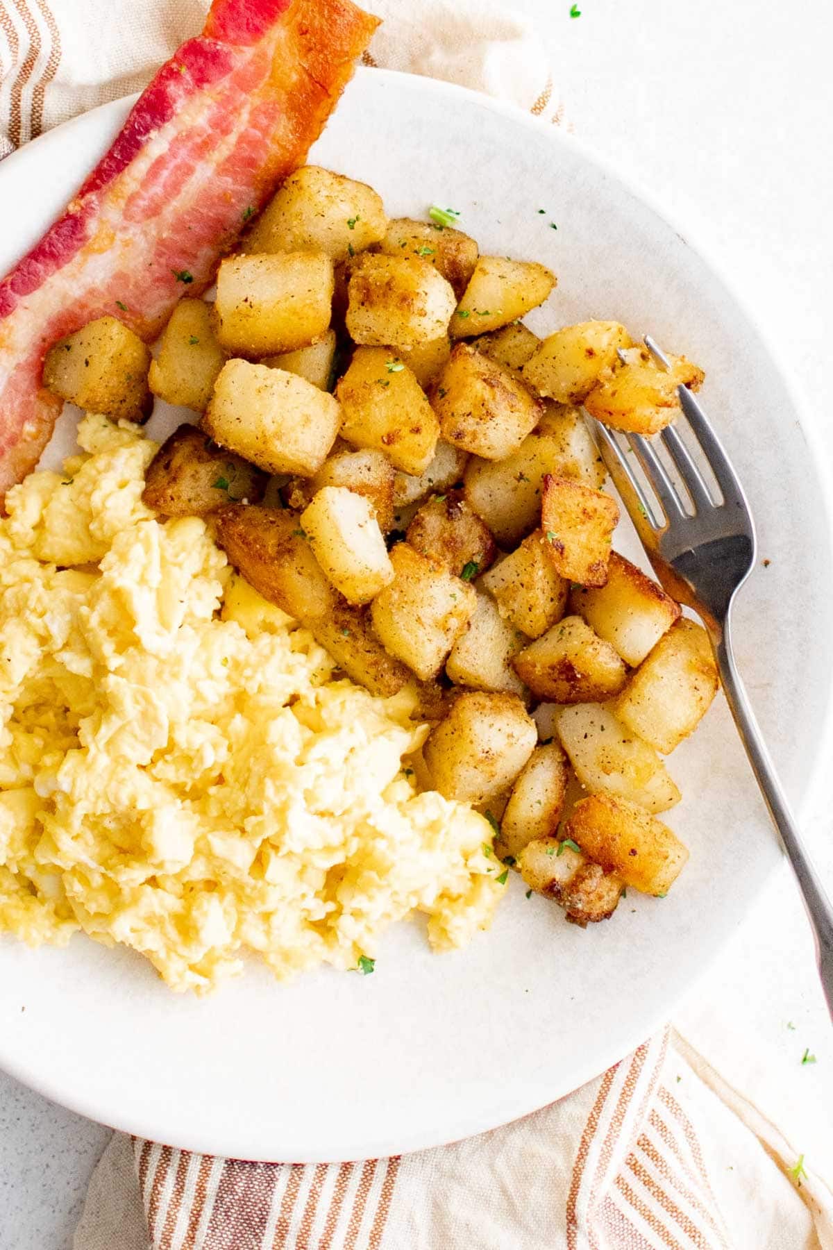 Crispy diced potatoes, bacon and scrambled eggs on a white plate with a fork.