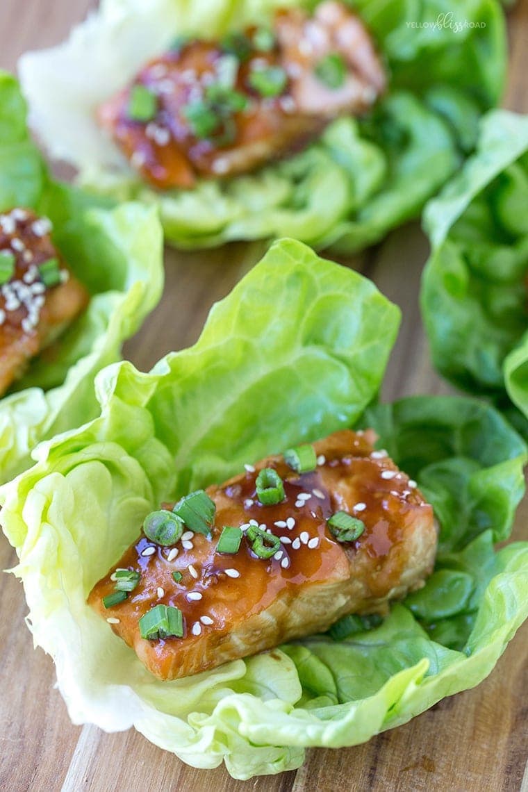 A wood cutting board with lettuce cups holding pieces of salmon with teriyaki sauce. They are garnished with sesame seeds and green onions.