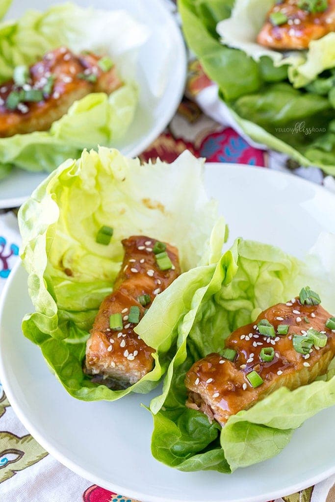 White plates with lettuce cups holding pieces of salmon with teriyaki sauce. They are garnished with sesame seeds and green onions.