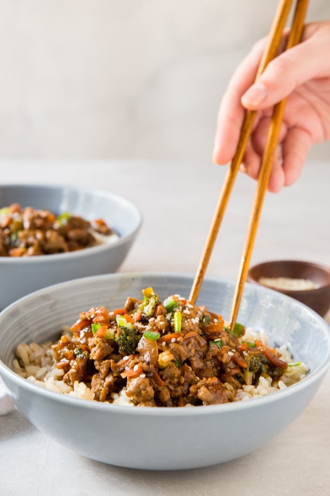 a hand with chopsticks, bowls with healthy ground turkey teriyaki, vegetables and rice