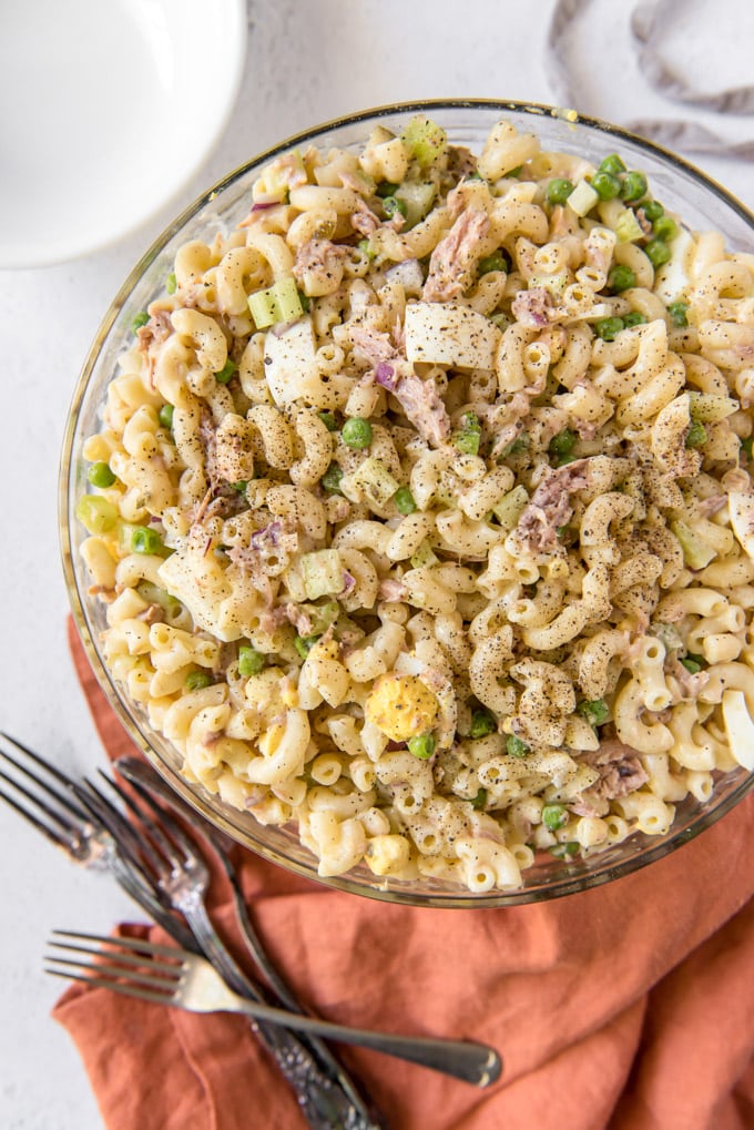 A large glass bowl filled with runa, macaroni, peas, celery and eggs. Forks and an orange napkin lay next to the bowl.